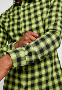 Groene en zwarte geruite flanellen overhemd met button-down kraag, gestructureerde stof en opgerolde lange mouwen die onderscheidende ruitpatronen tonen.