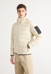 Man in een crèmekleurige, gewatteerde jas met capuchon, zwart label op de bovenste linkermouw, en een beige broek, staande tegen een effen achtergrond.