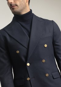 Blazer de doble botonadura en navy de una tela suave, con botones en tono dorado y solapa con muesca, combinado con un cuello alto a juego.