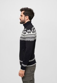 Suéter de punto en azul marino con un cuello alto, que presenta un patrón de copos de nieve blancos en los hombros y un acento geométrico blanco en el dobladillo.