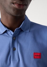 Gros plan d'une personne portant un polo bleu avec un écusson rouge étiqueté "HUGO" sur la poitrine et deux boutons défaits au col.