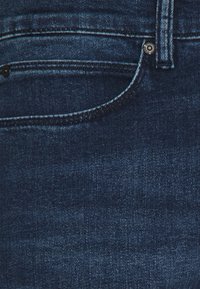 Tmavě modré džíny z denimu, detailní záběr na přední kapsu, švy, očka na opasek a kovové nýty.