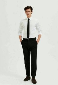 Balta krekls, melna kaklasaita, pielāgotas melnas bikses, brūnas kurpes. Gluda auduma, klasiskā piegriezuma, minimālisma dizains, stāvoša poza, rokas kabatās.