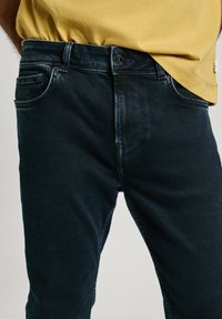 Dark blue denim jeans with a slim fit, featuring a front pocket, zip fly, and a subtle faded texture. Paired with a yellow cotton t-shirt.