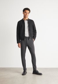 Veste en denim noir à fermeture à pression sur un t-shirt gris clair, associée à un pantalon gris foncé et des chaussures noires. Textures lisses et design ajusté.