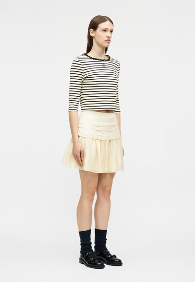 Γυναίκα όρθια, φοράει μαύρο και λευκό ριγέ cropped τοπ, κρεμ πολυεπίπεδη φούστα, μαύρες κάλτσες και μαύρα μοκασίνια, μπροστά σε απλό φόντο.