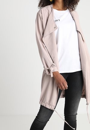 Femme portant un trench-coat beige clair sur un T-shirt blanc et un jean skinny noir, debout avec une main détendue le long du corps.