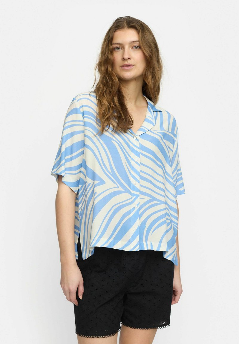 Camicia a maniche corte, con bottoni, di colore azzurro chiaro e bianco con un motivo a righe ondulate. Realizzata in tessuto morbido. Abbinata a pantaloni corti neri.