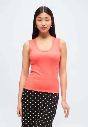 Hosszú fekete hajú nő korallos ujjatlan felsőt és fekete, fehér pöttyös szoknyát visel, egy egyszínű világos háttér előtt áll.