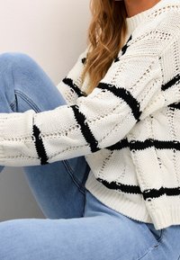 Pull en maille blanche avec des rayures noires, présentant un design texturé et un ourlet côtelé. Associé à un jean en denim bleu clair.