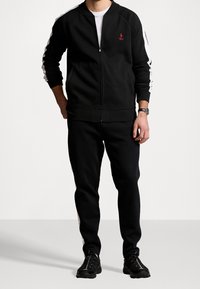 Černá sportovní tepláková souprava s bílými pruhy na stranách, bundou na zip, žebernými manžetami a kalhotami. Na hrudi je malá červená logo.