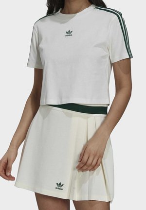 Camiseta corta blanca con logo de Adidas verde y rayas verdes en las mangas, combinada con una falda plisada crema que presenta un logo verde.