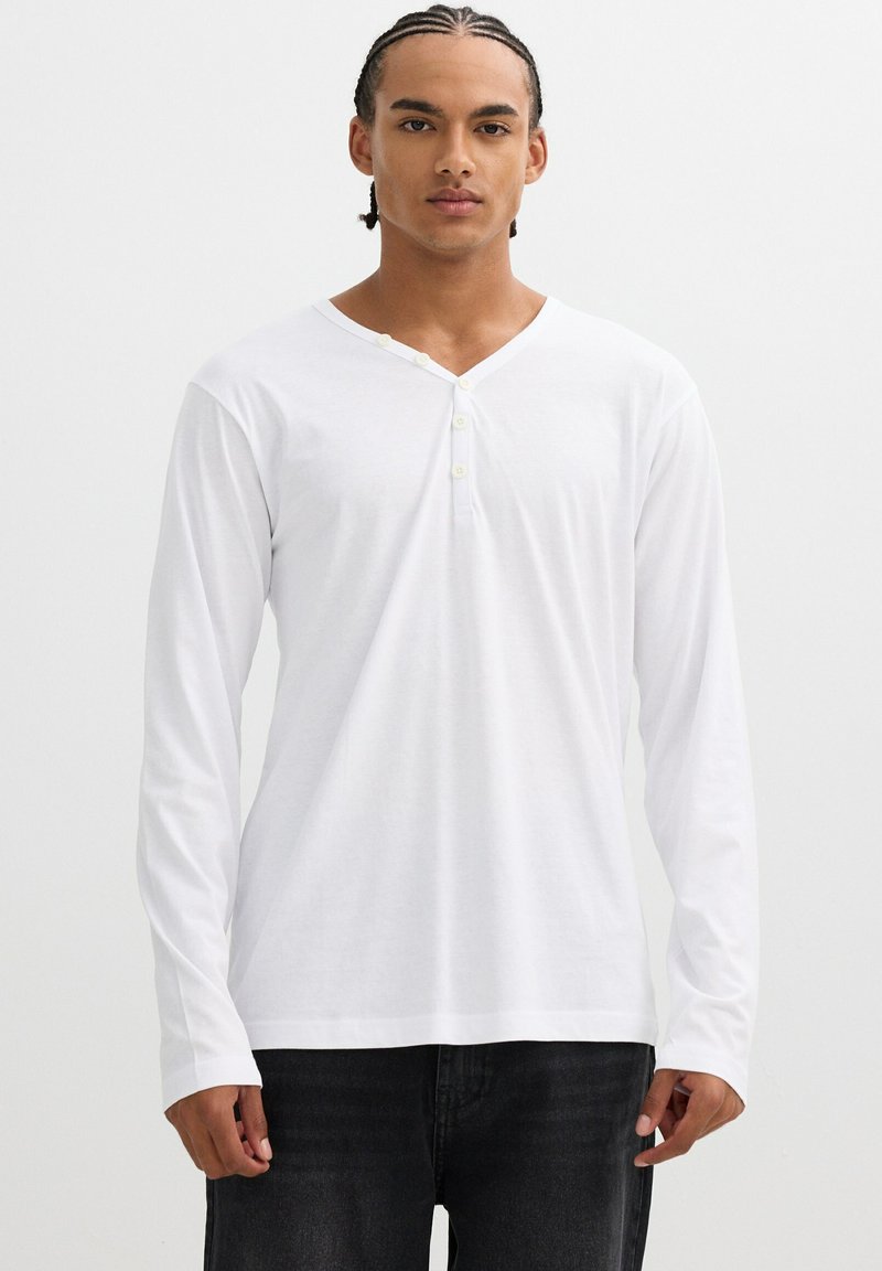 Camisa blanca de manga larga en tejido suave, con escote en V y tapeta de tres botones. Presenta un corte relajado y un diseño minimalista.