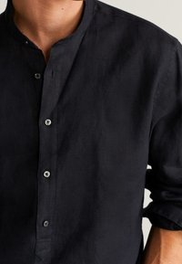 Uomo che indossa una camicia nera a maniche lunghe con colletto a band e quattro bottoni visibili, maniche arrotolate fino agli avambracci.