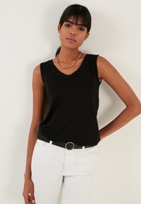 Black sleeveless top with a V-neck, made of soft fabric; paired with white pants and a black belt featuring a circular metal buckle.