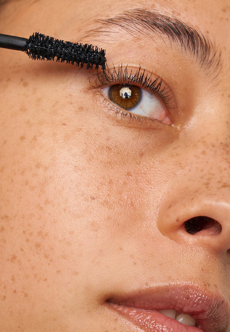 Brosse de mascara noire appliquant le produit sur de longs cils foncés. Gros plan sur une peau avec des taches de rousseur, un œil marron avec un reflet de lumière, et des lèvres lisses.