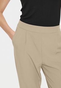 Pantalons beiges sur mesure avec une texture lisse et des plis, dotés de poches latérales. Porté avec un haut noir, mettant en valeur un design épuré.