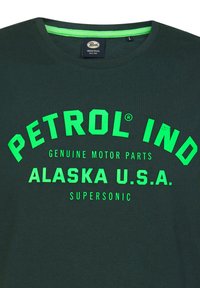 Tmavě zelené bavlněné tričko s jasně zeleným potiskem: "PETROL IND," "ALASKA U.S.A.," a "SUPERSOIC." Kulatý výstřih, krátké rukávy.