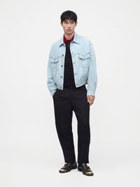 Jeune homme debout portant une veste en jean bleu clair, un polo noir à col rouge, un pantalon noir et des chaussures noires avec des bouts dorés.