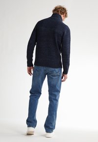 Pulover bleumarin cu guler înalt și textură canelată; asortat cu blugi albaștri și adidași albi. Vedere din spate, evidențiind designul tricotat.