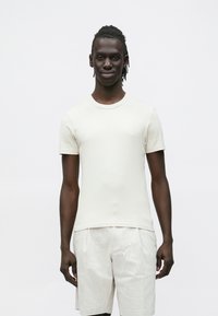 Jovem de pele escura e dreadlocks, vestindo uma camisa branca simples de manga curta e calções bege claros, em pé contra um fundo branco.