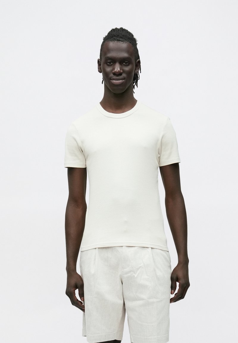 Jovem de pele escura e dreadlocks, vestindo uma camisa branca simples de manga curta e calções bege claros, em pé contra um fundo branco.