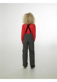 Grijze overalls met zwarte banden over een rode blouse, met een relaxte pasvorm en zijzakken. Achteraanzicht toont krullend haar, verder geen aanvullende details.
