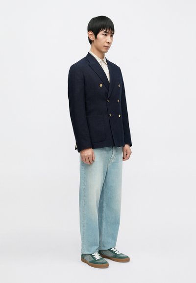 Granatowa marynarka dwurzędowa ze złotymi guzikami, jasna koszula, jasnoniebieskie dżinsy i zielone trampki z białymi sznurowadłami na neutralnym tle.