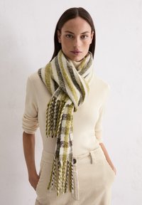 Écharpe rayée aux teintes vert, blanc et marron, présentant une texture douce et des extrémités à franges. Portée sur une tenue crème.