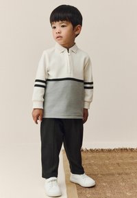 Langärmliges Polo in weißem und grauem strukturiertem Stoff mit schwarzen Streifen, kombiniert mit dunklen Hosen und weißen Sneakers, vor einem neutralen Hintergrund.