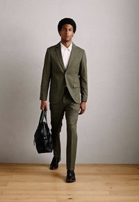 Hombre caminando por dentro, vestido con un traje verde oliva y una camisa blanca, llevando un bolso de cuero negro en su mano izquierda.