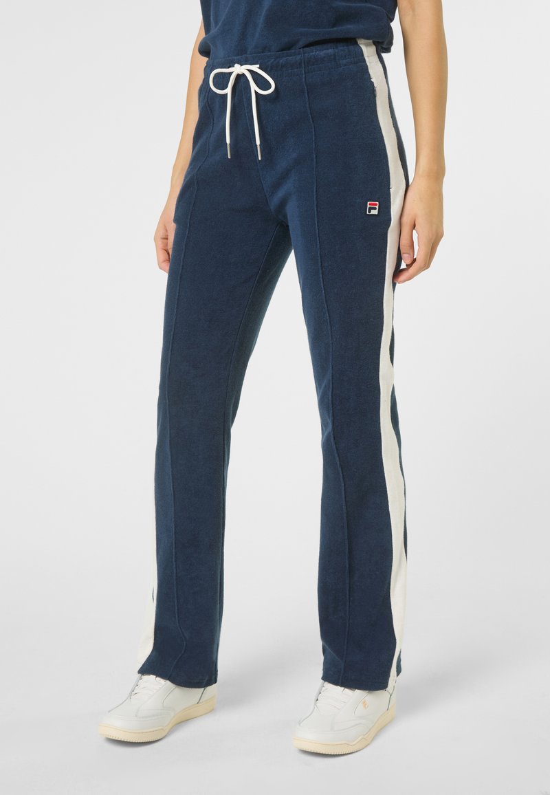 Osoba nosząca ciemnoniebieskie spodnie z białymi paskami po bokach, białym sznurkiem w talii i białe trampki na jasnym tle.