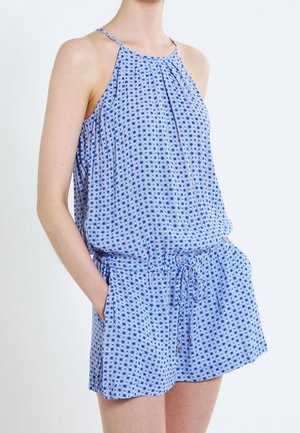 Frau trägt einen blauen ärmellosen Overall mit weißem Blumenmuster und Kordelzug in der Taille, Hände in den Taschen des Overalls.