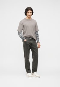 Νεαρός άνδρας όρθιος, φοράει γκρι κοντομάνικο πουκάμισο πάνω από μπλούζα με μοτίβο ζέβρας, σκούρα τζιν με ζώνη με κρίκους και άσπρα αθλητικά παπούτσια.