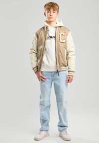 Beige varsity-takki vaaleilla hihansuilla, rinta  iso "C", valkoinen huppari tekstillä, vaaleansiniset farkut ja valkoiset lenkkarit.
