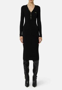 Vestito nero in maglia con maniche lunghe, scollo a V e bottoni dorati. Silhouette aderente e lunghezza al ginocchio, abbinato a stivali neri lunghi.