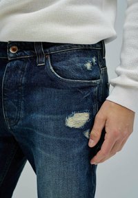 Vaqueros de denim azul desgastado con parches deshilachados. Presentan un corte ajustado, lavado oscuro y bolsillos estándar. Mano posicionada en la cadera.