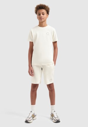 Tienerjongen staat met handen in zijn zakken, draagt een crèmekleurig t-shirt en korte broek met sneakers, tegen een effen lichte achtergrond.