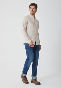 Lyse beige linned skjorte med ståkrave, parret med blå denimjeans og brune læderstøvler, med oprebne ærmer.