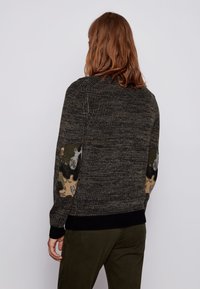 Stickad tröja i mörka nyanser med en texturerad kropp och kamouflage-mönstrade armbågsplåster. Ribbad fåll och ärmslut med svarta accenter.