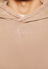 Gros plan d'un sweat à capuche beige avec une broderie blanche portant l'inscription « Kani » sur la poitrine, porté par-dessus une chemise blanche.