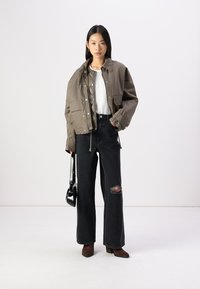 Giacca verde oliva tagliata con bottoni a pressione, abbinata a jeans neri a gamba larga con strappi sulle ginocchia. Stivali alla caviglia marroni e una borsa nera completano il look.