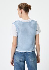 Chaleco de punto azul claro con un patrón texturizado, de manga corta, llevado sobre una camiseta blanca, combinado con unos jeans de mezclilla azul de tiro alto.