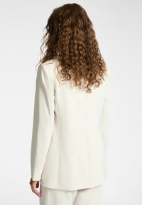 Mujer con cabello rizado largo vestida con un blazer ajustado color blanco roto y pantalones a juego, de espaldas frente a un fondo liso.