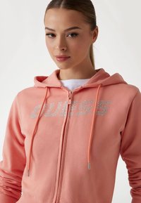 Haut à zip rose avec capuche, arborant un logo "GUESS" scintillant argenté, des poignets côtelés et des cordons de serrage avec des accents argentés.