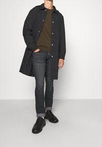 Hombre con abrigo gris oscuro abrochado, suéter verde oliva, vaqueros negros con puños, calcetines blancos y zapatillas negras, de pie con una mano en el bolsillo.