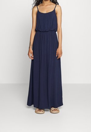 Vrouw draagt een marineblauwe maxi-jurk zonder mouwen met een geplooide taille en beige sandaal met touw, staand tegen een effen achtergrond.