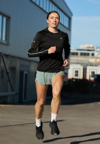 Crna dugouha sportsku majicu s bijelim detaljima, mint zelene kratke hlače za trčanje i crne tenisice za trčanje. Trkač u opuštenom vanjskom okruženju.