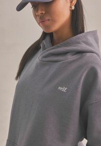 Szary, zakapturony sweatshirt o miękkiej fakturze, z subtelnym haftowanym logo "self." na piersi, noszony z dopasowaną czapką.