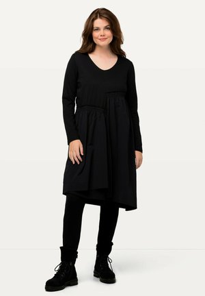 Schwarzes, langärmliges Kleid mit V-Ausschnitt und gerafftem Taillenbereich, mit einem unebenen Saum, kombiniert mit schwarzen Leggings und Schnürstiefeln.